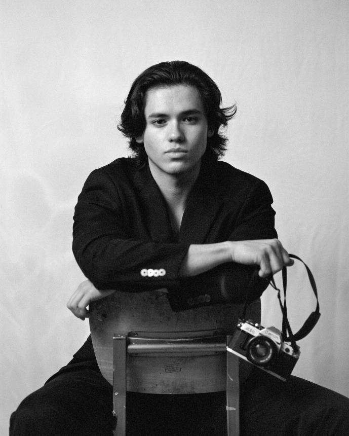 Alex Hoefer sits straddling a chair, holding an analog camera