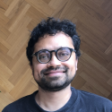 Headshot photo of speaker Dr. Siddique standing in front of a geometric pattern wood background. He is wearing a dark shirt, glasses, and smiling.