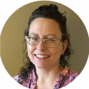 A photo of Dr. Jill Bradbury smiling. She has fair skin and curly hair, and is wearing glasses, dangly earrings, and a patterned top